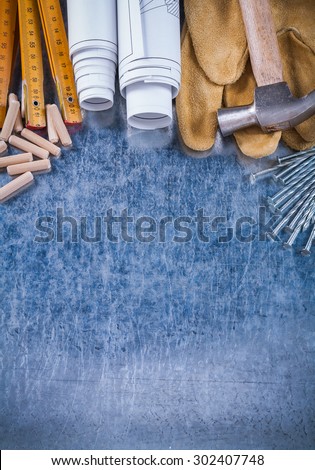 Blueprint rolls claw hammer safety gloves wooden meter steel nails and dowels on scratched metallic background construction concept.
