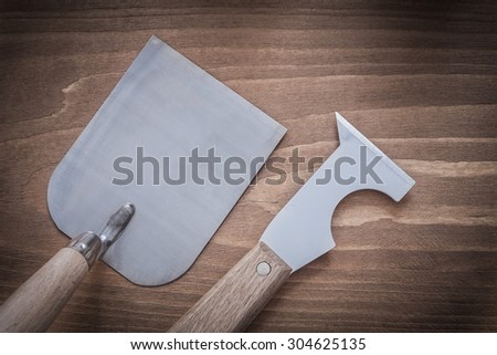 Sharp glossy paint scraper and bricklaying trowel on wooden surface.