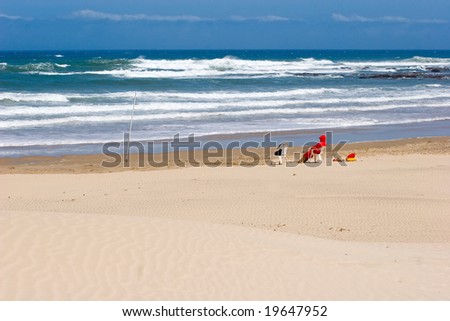 lifeguard with red jacket on-duty at an empty beach with sand,ocean and sky visible,seated on white plastic chair