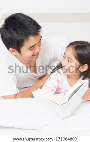 Little Asian child reading a story book with father on the bed
