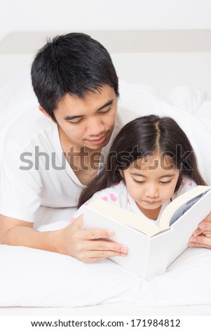 Little Asian child reading a story book with father on the bed