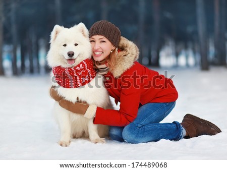 Joyful beautiful stylishly dressed young woman in red jacket hugging white Samoyed dog outdoors in winter day