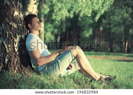 Relaxing man sitting near a tree with his eyes closed meditating and enjoying the warm summer sunset