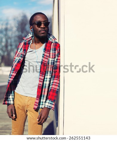 Outdoor fashion portrait of stylish young african man listens to music and enjoys freedom, wearing a plaid hipster red shirt and sunglasses, posing near urban white wall for background, copy space
