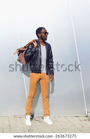 Outdoor fashion portrait of handsome african man in black leather jacket with bag standing against the shiny metal wall