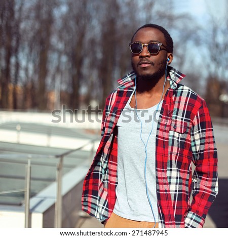 Fashion portrait of stylish young african man listens to music in earphones outdoors, handsome hipster wearing a plaid red shirt and sunglasses