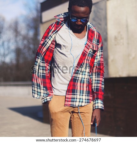 Street fashion concept - portrait of stylish young african man standing listens to music in earphones outdoors, handsome hipster wearing a plaid red shirt and sunglasses