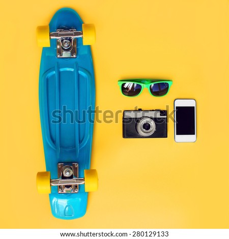 Fashion summer look concept. Blue skateboard, green sunglasses, vintage camera and screen smartphone on a yellow background, top view. Trendy colorful photo