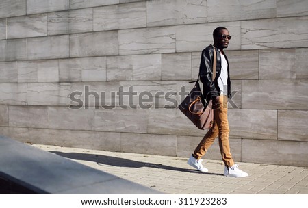 Fashion stylish young african man in sunglasses and black rock leather jacket with bag walking over grey textured city background