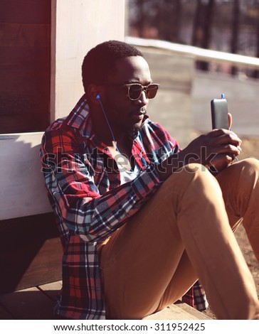 Lifestyle fashion portrait of stylish young african man listens to music and using smartphone, wearing a hipster plaid red shirt and sunglasses sitting in the city evening