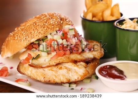 Delicious chicken burger with chopped salad served with potato wedges on a white plate on a wooden table.