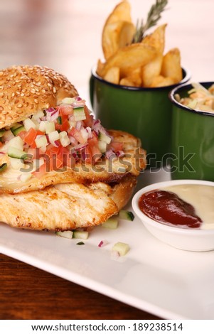 Delicious chicken burger with chopped salad served with potato wedges on a white plate on a wooden table.