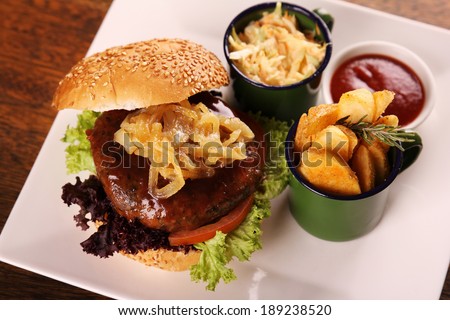 Delicious juicy beef burger with caramelised onions and salad served with potato wedges on a white plate on a wooden table.