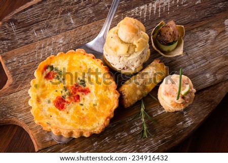A variety of delicious savory pastries and bites served on a wooden board. The selection includes quiches, spring rolls, scones with chicken and seafood.
