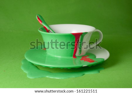 White cup with a saucer and a spoon poured with red and green paint on a green background