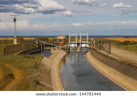 Gates in channel or irrigation dam with electric control booth. Sunny landscape, flat and dry with wind turbines (windmills) in the background