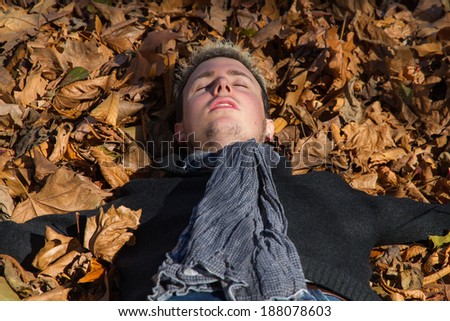 Young man in the park, with eyes closed and arms outstretched, lying in the sun on dry autumn leaves