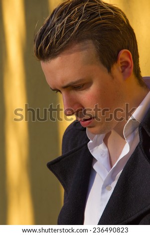 Young man looking down, with jacket, in front of an illuminated with warm rays of sunset wall