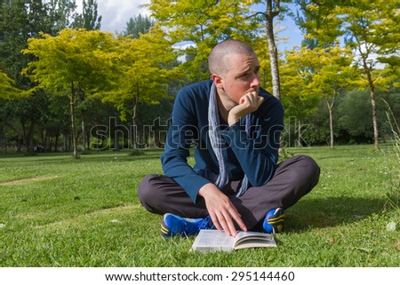Young man with cropped hair sitting on the grass in a park and cross legged with a book in hand, looking around dispersed