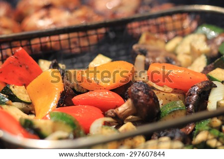 Grilled vegetables including zucchini, mushrooms, onions and peppers in a grill basket on a barbecue grill being cooked with chicken in background