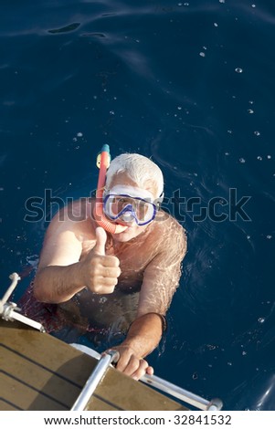 The happy senior man swim in the blue sea with his thumb up.