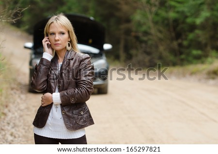 Young beautiful sexy woman at broken car with mobile phone, standing in the public road in forest area, calling for help with mobile phone. Broken vehicle in the background.