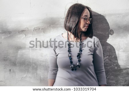 Plus size model with bob haircut wearing minimalist gray maxi dress, sunglasses and jewelry posing over street wall.