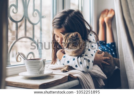 Child in pajamas relaxing on a window sill with pet. Lazy weekend with cat at home. Cozy scene, hygge concept.