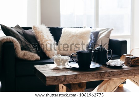 Still life details of nordic living room. Cup of hot tea with steam on a rustic coffee table over black sofa in morning sunlight. Cozy winter concept in scandinavian home interior.
