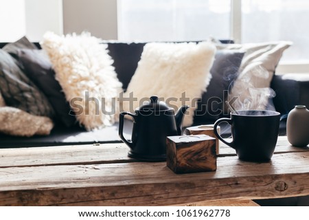 Still life details of nordic living room. Cup of hot tea with steam on a rustic coffee table over black sofa in morning sunlight. Cozy winter concept in scandinavian home interior.