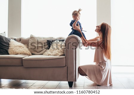 Mom with daughter playing in beautiful holiday dresses on sofa in luxury interior. Baby princess fashion look.