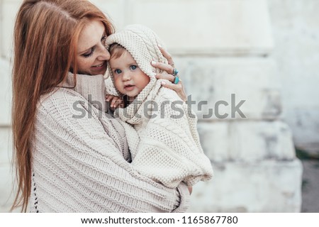 Portrait of beautiful mom holding on hands her 8 months old baby. Both dressed in warm winter knitted clothing.
