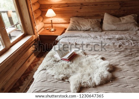 Rustic interior of log cabin bedroom with bed by big window. Opened book on sheep rug. Warm and cozy weekend morning in hotel.