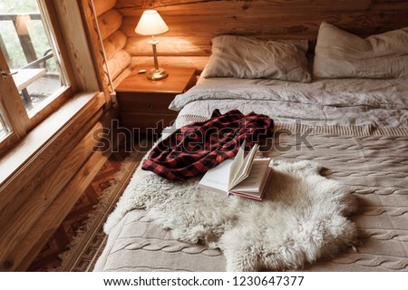 Rustic interior of log cabin bedroom with bed by big window. Opened book on sheep rug. Warm and cozy weekend morning in hotel.
