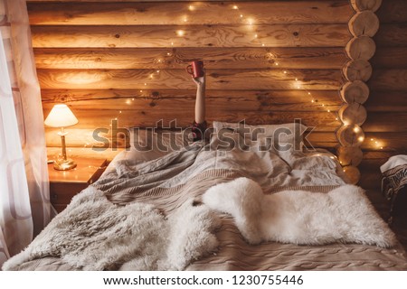 Woman relaxing and drinking morning coffee on cozy bed in log cabin in winter