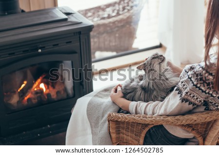 Human with cat relaxing in wicker armchair by the fire place in wooden cabin. Warm and cozy winter holiday concept.