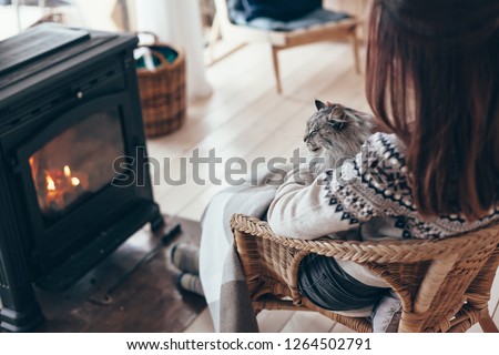 Human with cat relaxing in wicker armchair by the fire place in wooden cabin. Warm and cozy winter holiday concept.
