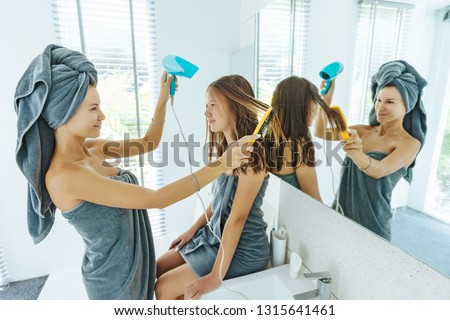 Mom with teenage dauhter making everyday routine in modern hotel bathroom together. Mother is brushing and drying child hair after shower.