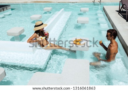 Teenage children relaxing while eating pizza and drinking cocktail in the hotel pool. Luxury tropical beach lifestyle.