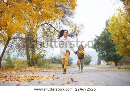Young mother with her little daughter walking in fall park on yellow fallen leaves one autumn day