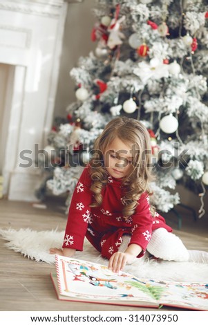 5 years old little girl reading book with winter tales from Santa Claus near Christmas tree in morning at home