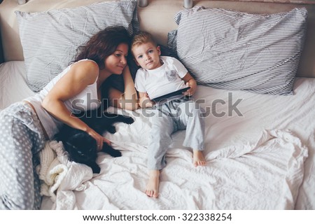 Young mother with her 2 years old little son dressed in pajamas are relaxing and playing in the bed at the weekend together, lazy morning, warm and cozy scene. Pastel colors, selective focus.