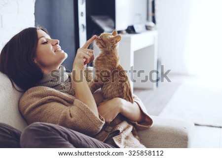 Young woman wearing warm sweater is resting with a cat on the armchair at home one autumn day