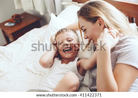 Young mother with her 6 years old little son dressed in pajamas are relaxing and playing in the bed at the weekend together, lazy morning, warm and cozy scene.