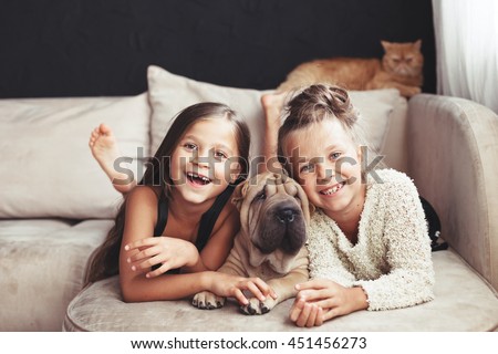 Home portrait of two cute children hugging with ginger cat and puppy of Chinese Shar Pei dog on the sofa against black wall