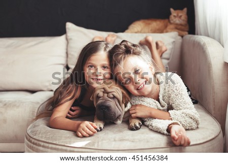 Home portrait of two cute children hugging with ginger cat and puppy of Chinese Shar Pei dog on the sofa against black wall