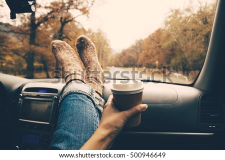 Woman feet in warm socks on car dashboard. Drinking take away coffee on road. Fall trip. Rain drops on windshield. Freedom travel concept. Autumn weekend.