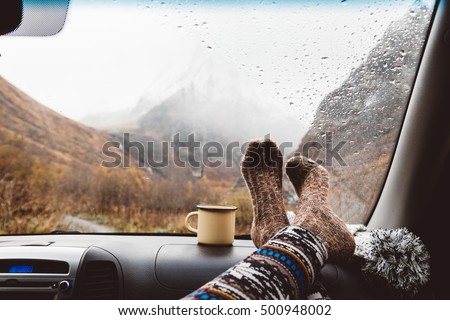 Woman legs in warm socks on car dashboard. Drinking warm tee on the way. Fall trip. Rain drops on windshield. Freedom travel concept. Autumn weekend in mountains.