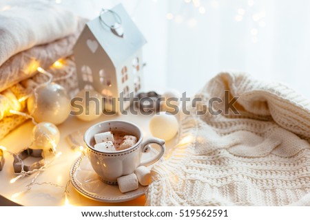 White Christmas decor: warm sweater, cup of hot cocoa with marshmallow and led string lights. Winter mood, holiday decoration.