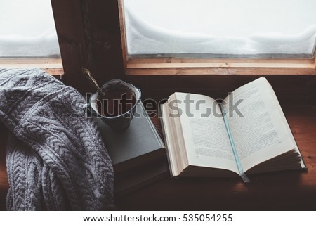 Warm and comfy winter concept. Book, cup of lemon tea and sweater on wooden window sill in old house. Reading and relaxing in cold snowy weather at home. Quiet silent homely scene.